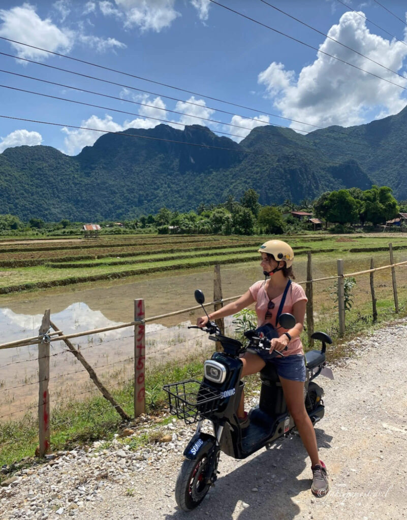 Guada sits on an e-book and looks out at the lush landscape. The background has lots of greens, and some water in the rice fields is reflecting the rock formations behind