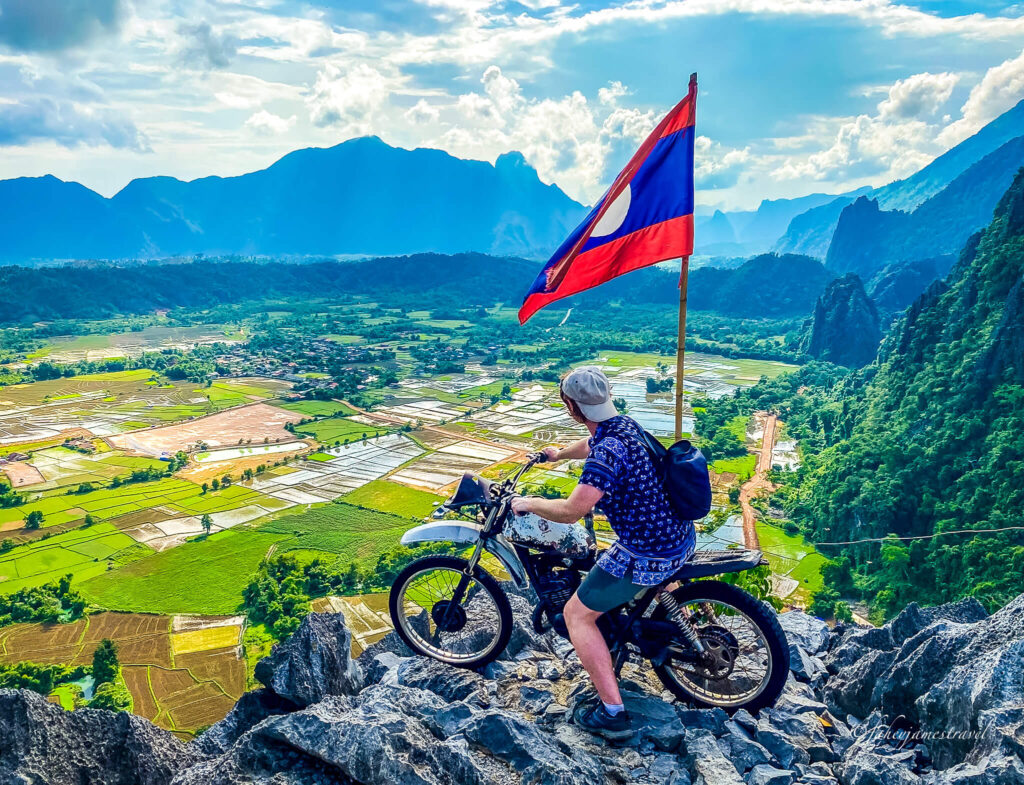 james sits on the bike at Pha Ngern viewpoint in Vang Vieng, where the Laos flag is blowing in the wind above his head. The rice fields are green and lush
