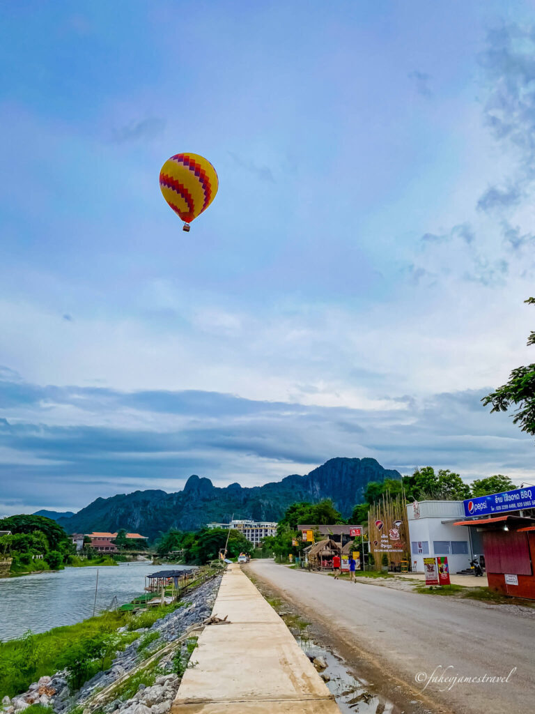 A hot air balloon is shown high in the sky over Vang vieng. The river is to the left of the image, and the dome shop is to the right. A couple of people walk away from the camera on the road.