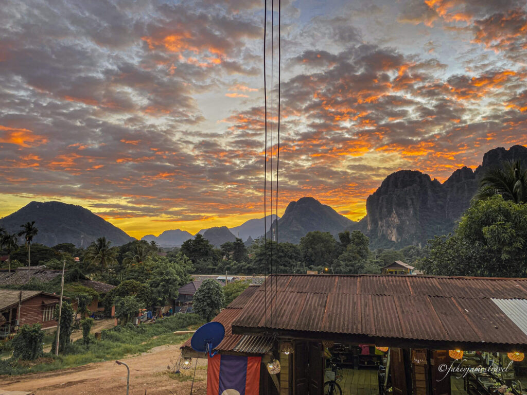 sun sets over the karst limestone formations in vang vieng. a shed is in the front of the picture, and the sky is cloudy and red, and orange