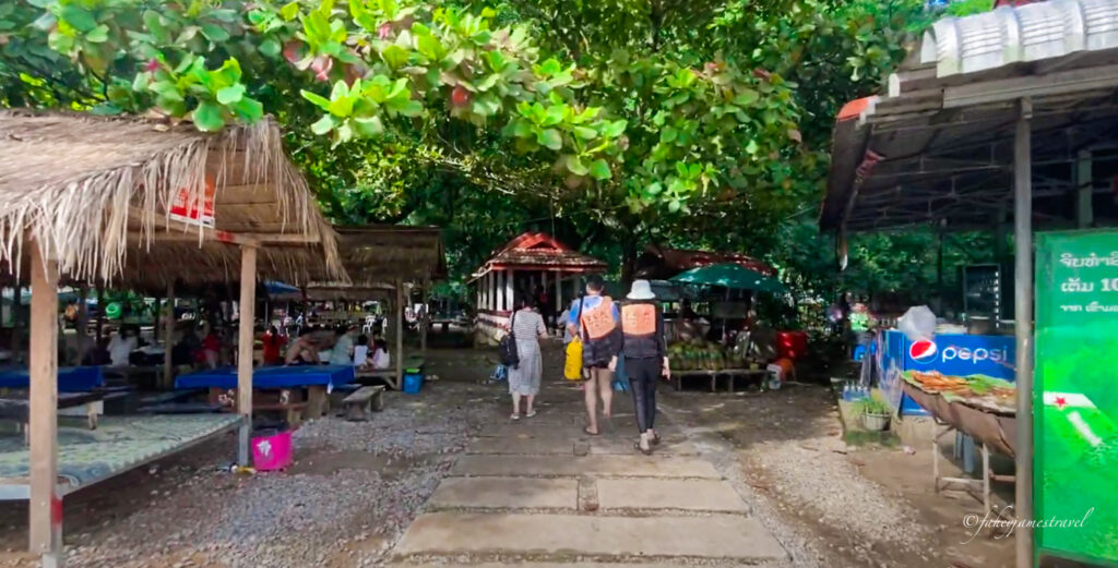 2 people are shown walking into the blue lagoon 1 while there are shops on either side to buy drinks and snacks. Green trees are growing overhead