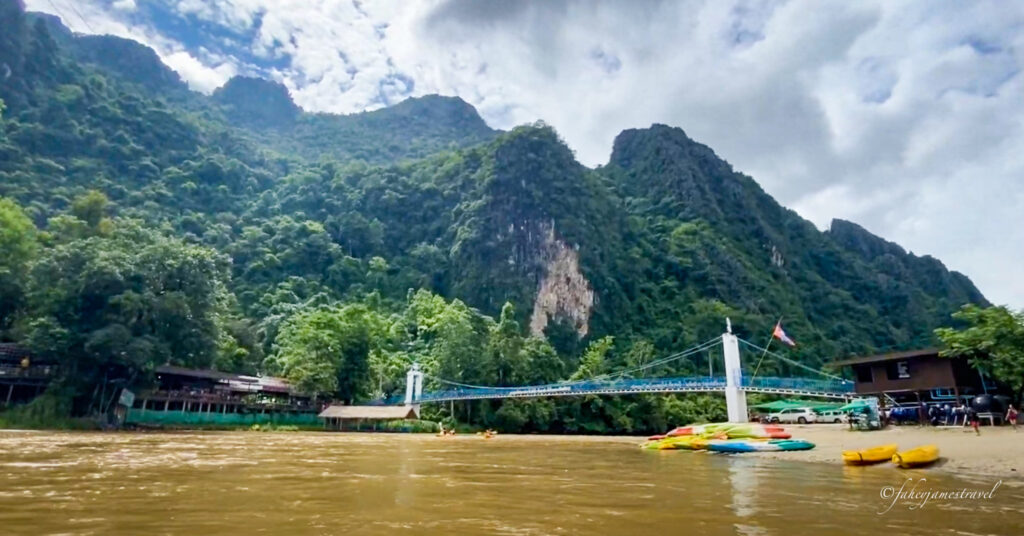 An epic shot of the limestone formations in Vang Vieng. There is a blue bridge across the river, which looks small compared to the formations. There is a lot of greenery, and the river is a muddy colour