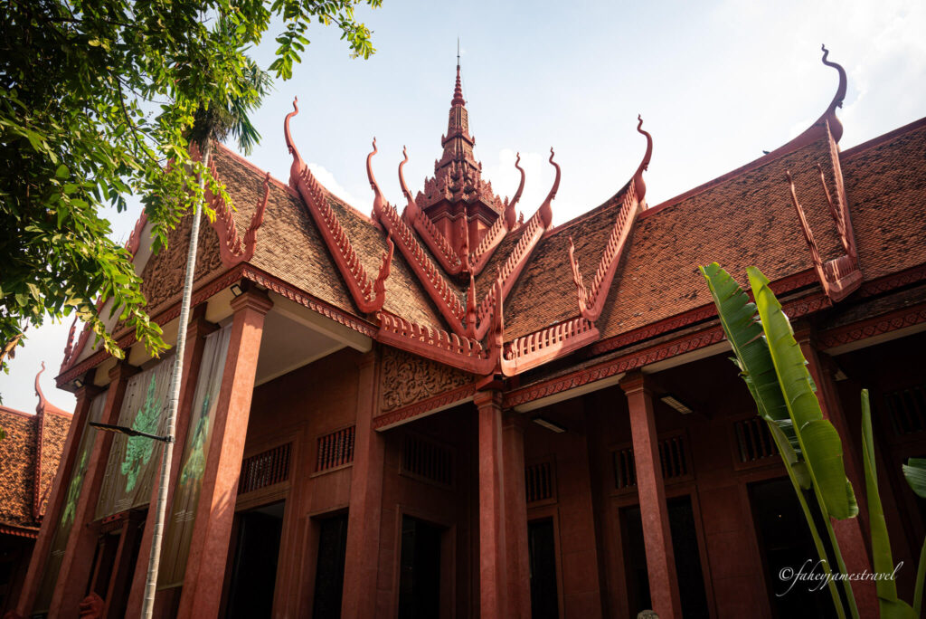 The exterior of the National Museum of Cambodia in Phnom Penh is shown. It has a red terracotta design with some trees on the sides. 