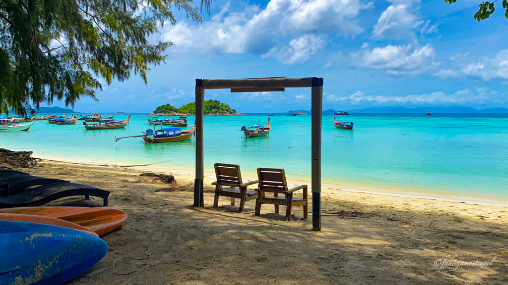 two seats si under a sign post on sunrise beach on koh lipe with some longtail boats sitting in the calm water. the water is blue and looks tropical