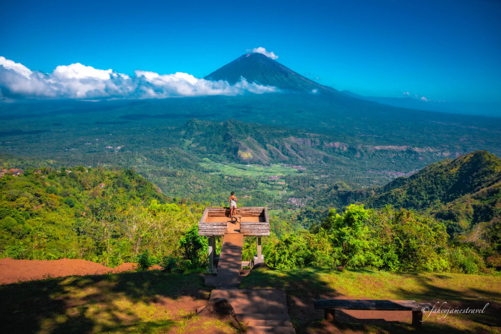 guads stands at one of the viewpoint made from woof and is elevated up. in the background you can clearly see mount agung and some cloud below the summit
