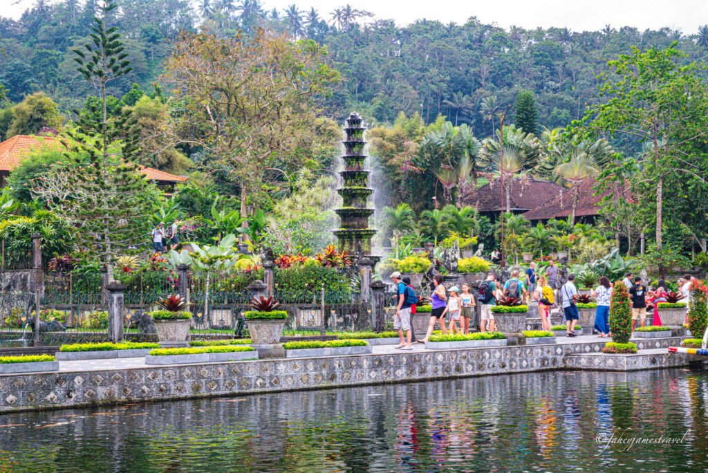 Tirta Gangga water palce has amazing detail. a fountain is pouring water from the top and falls lower with many people around it in the centre of the screen 