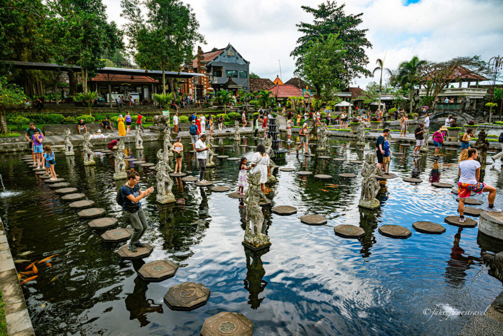 Tirta Gangga has a pond with many stepping stones where poeple are pictured standing. there are some green trees and the sky is cloudy