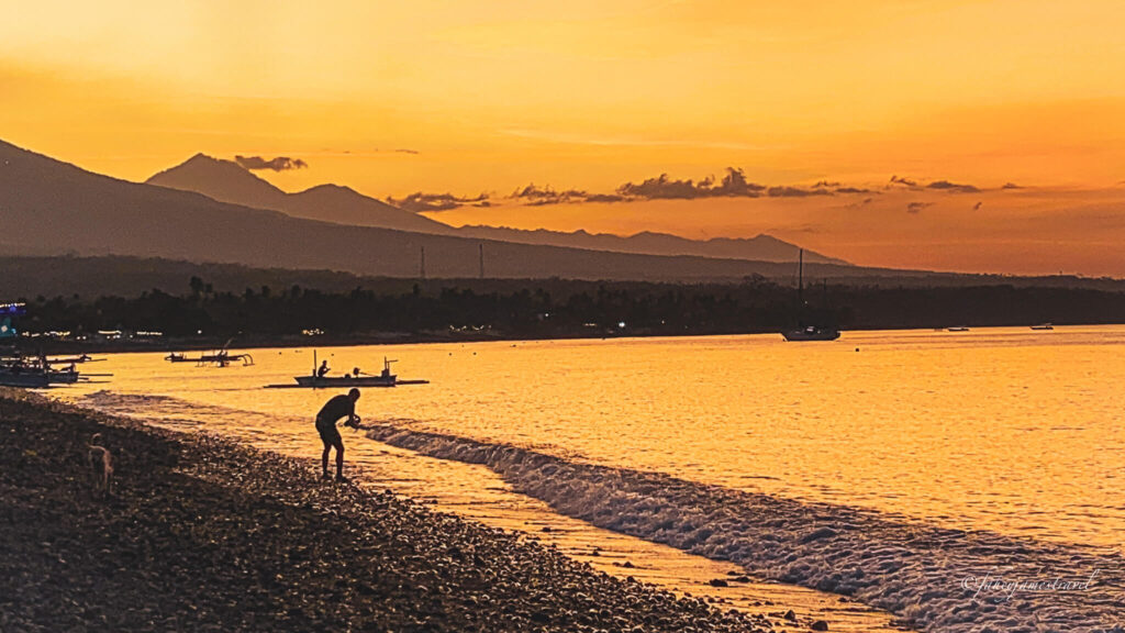a man stands beside the shore on Pantai Amed as the sun sets the sky is yellow and the sea is calm
