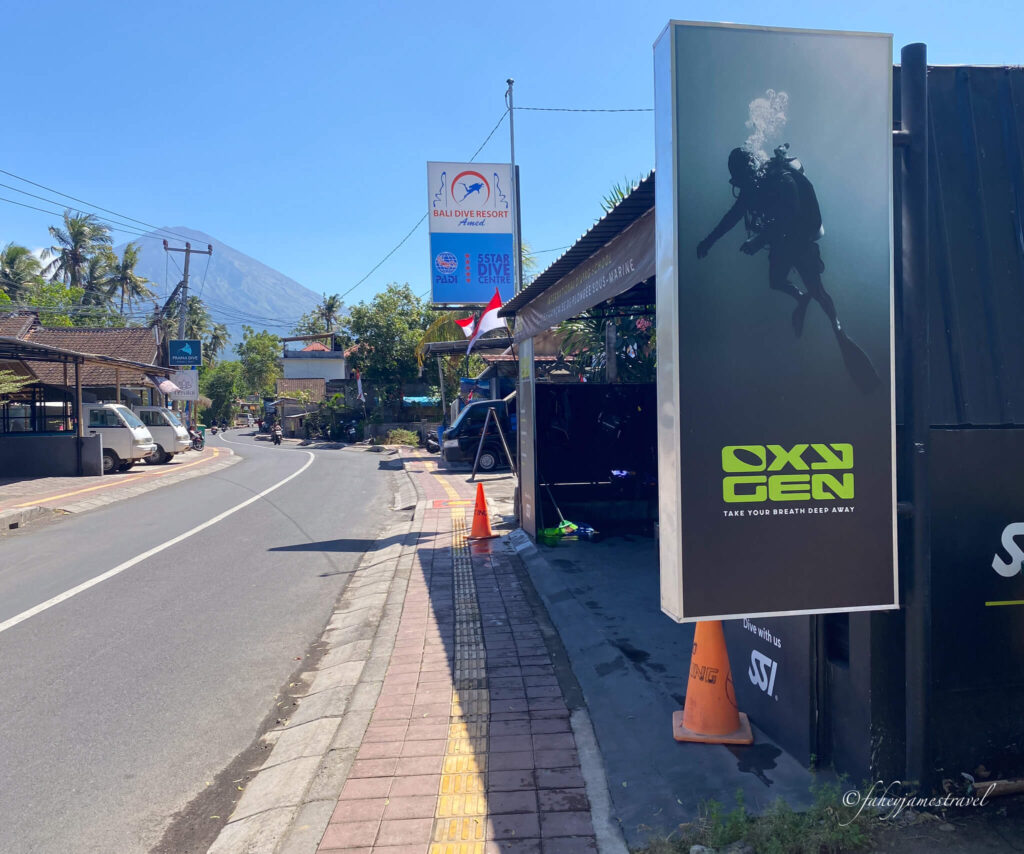 oxegen dive centre close to amed beach. the road is right beside the store with no traffic on the road. the sky is clear and you can see mount agung only barely down the road