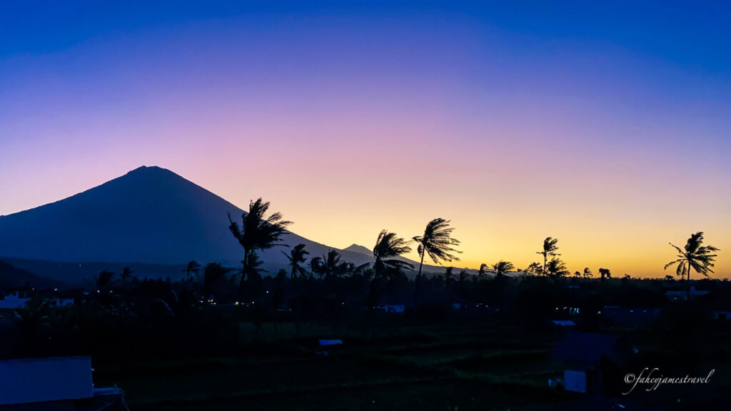 mount agung stands in the background with some palm trees blowing in the wind as the sun sets. the sky is blue and yellow 