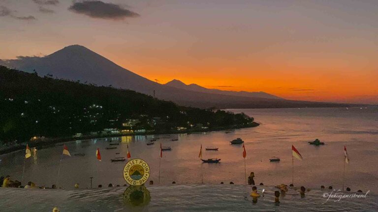 a man stands on pantai amed as the sun sets over mount agung, the sky is yellow with some pink