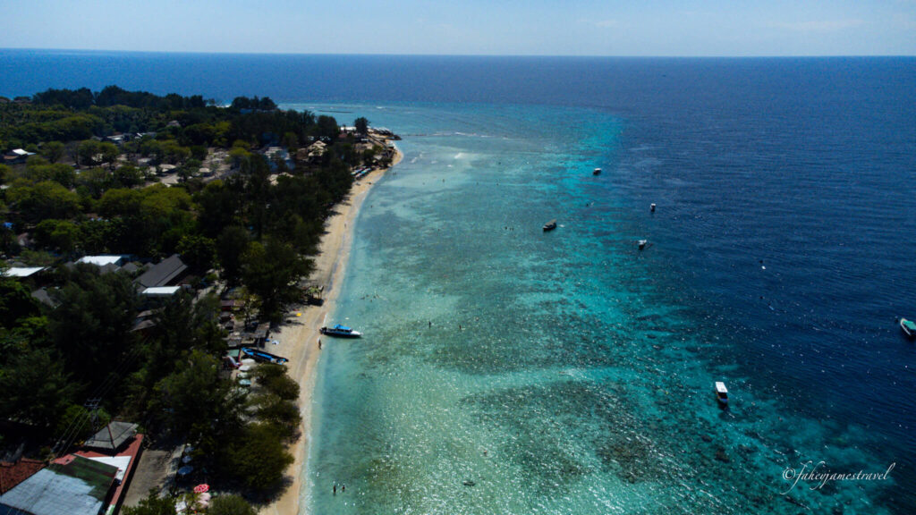 a long stretch of beach on gili t. the left side of the image is dark green with treets and the middle is light blue water and the right is darker blue