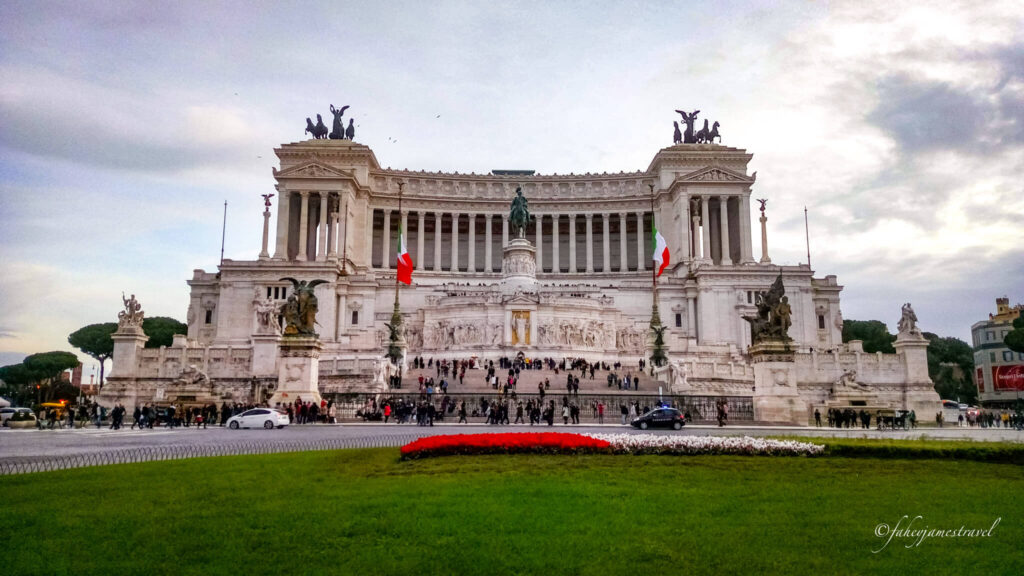 the alter of the fatherland. some gass is visible in the foregrouns and the sky has cloud. the building is white marble 