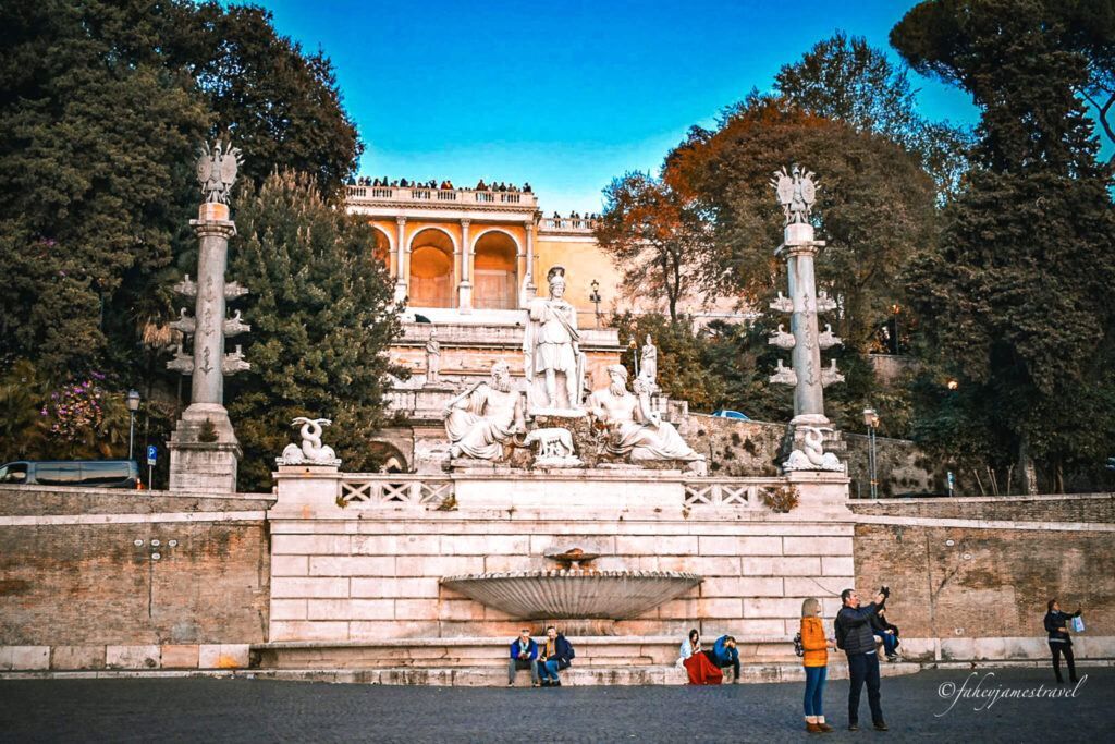 a couple stand for a photo inside Piazza del Popolo while people are visible on the terrace above 