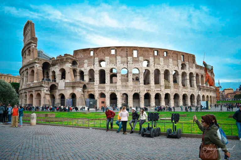 a shot of the colosseum from outside. the sky is blue and people walk past