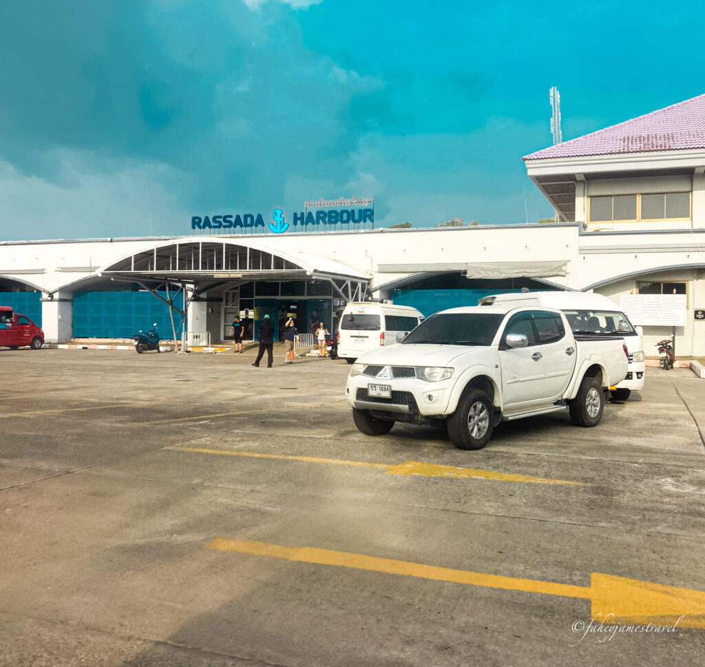 Rassada Pier is shwon with one white private taxi on the right of the image and one woman standing waiting for a taxi in the background. the sky is blue with little cloud 